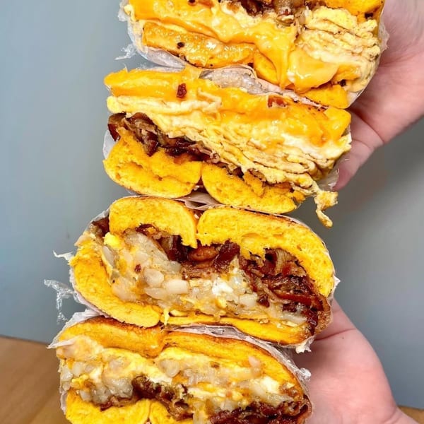 Two hands hold four halves of The Bagels' breakfast bagels, filled with eggs, cheese, and meat.