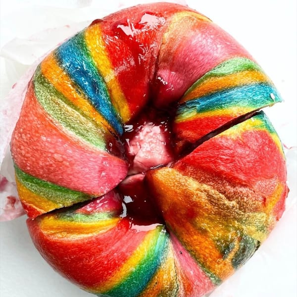 A vibrant rainbow bagel, sliced into quarters, filled with cream cheese and strawberry jam.