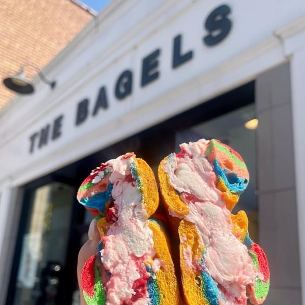 Rainbow bagel with pink cream cheese filling, held in front of The Bagels storefront.