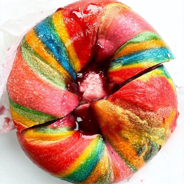A vibrant rainbow bagel from The Bagels, sliced and filled with a red jelly.