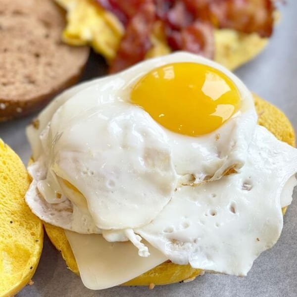 Close-up of a bagel with a fried egg, runny yolk, and melted cheese, with bacon in the background.