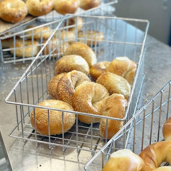 Freshly baked sesame bagels in metal baskets on a stainless steel counter at The Bagels.