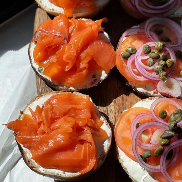 Four bagels on a wooden board, two with lox and two with tomato, red onion, and capers.