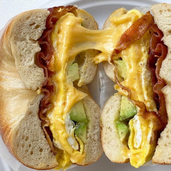 A bagel sandwich with bacon, melted cheese, scrambled eggs, and avocado, cut in half.