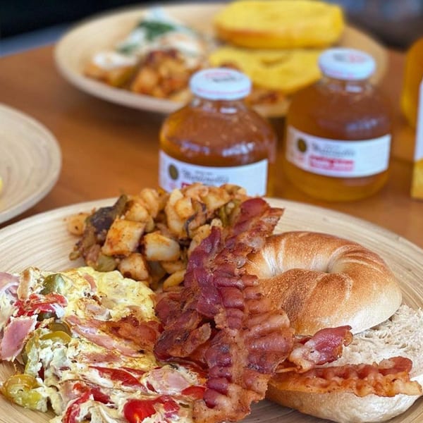 Breakfast plate with scrambled eggs, bacon, home fries, and a bagel, with bottled drinks in the background.