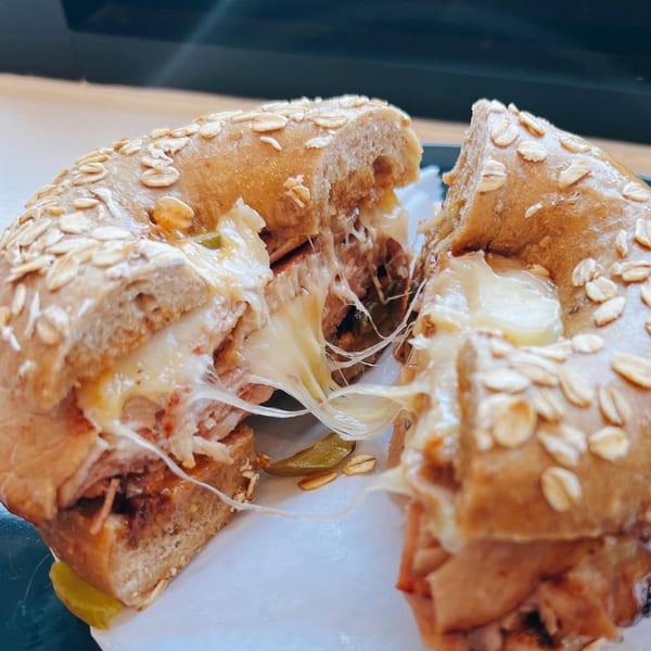 A sliced oatmeal bagel from The Bagels, filled with melted cheese and meat, stretching apart.